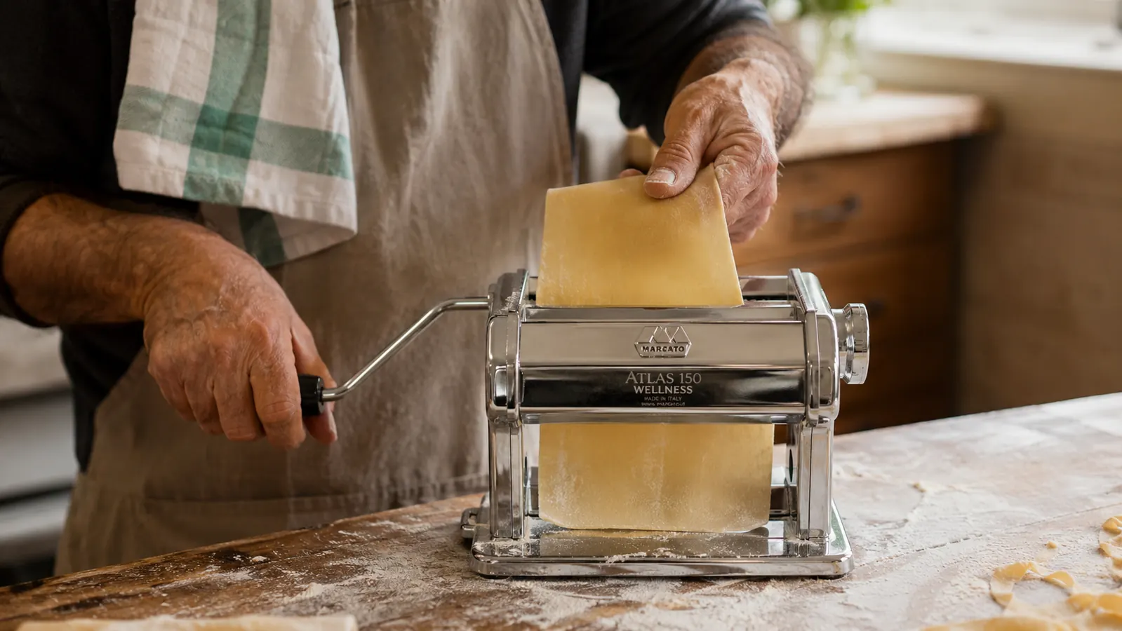 A hand turning the crank of the Marcato Atlas pasta machine clamped to a kitchen counter