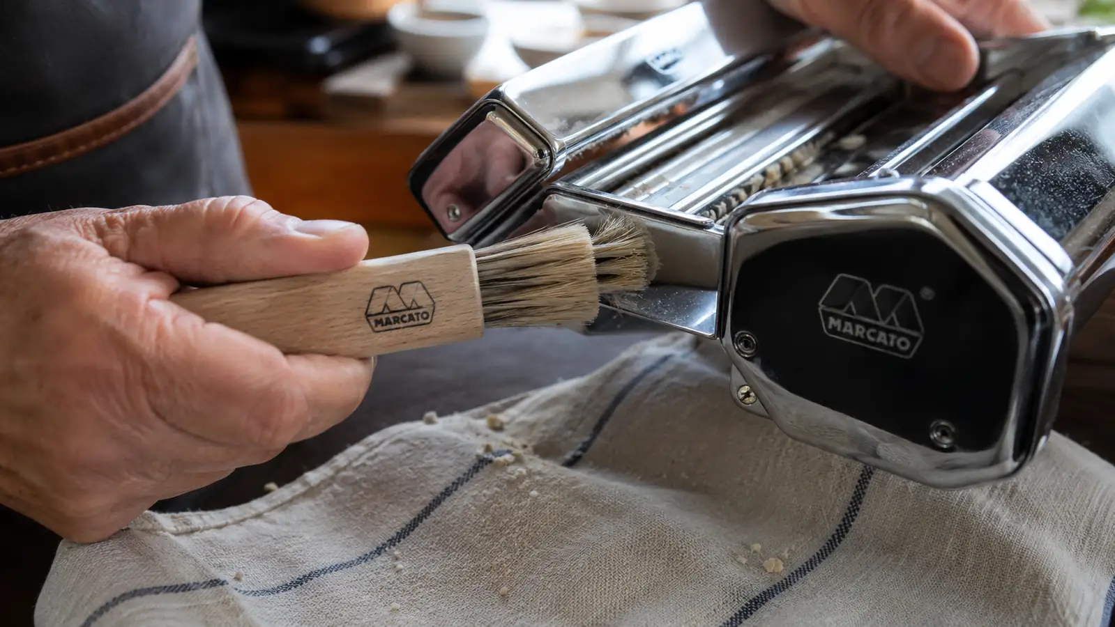 A stiff-bristled brush cleaning dried dough flecks out of pasta machine rollers on a towel