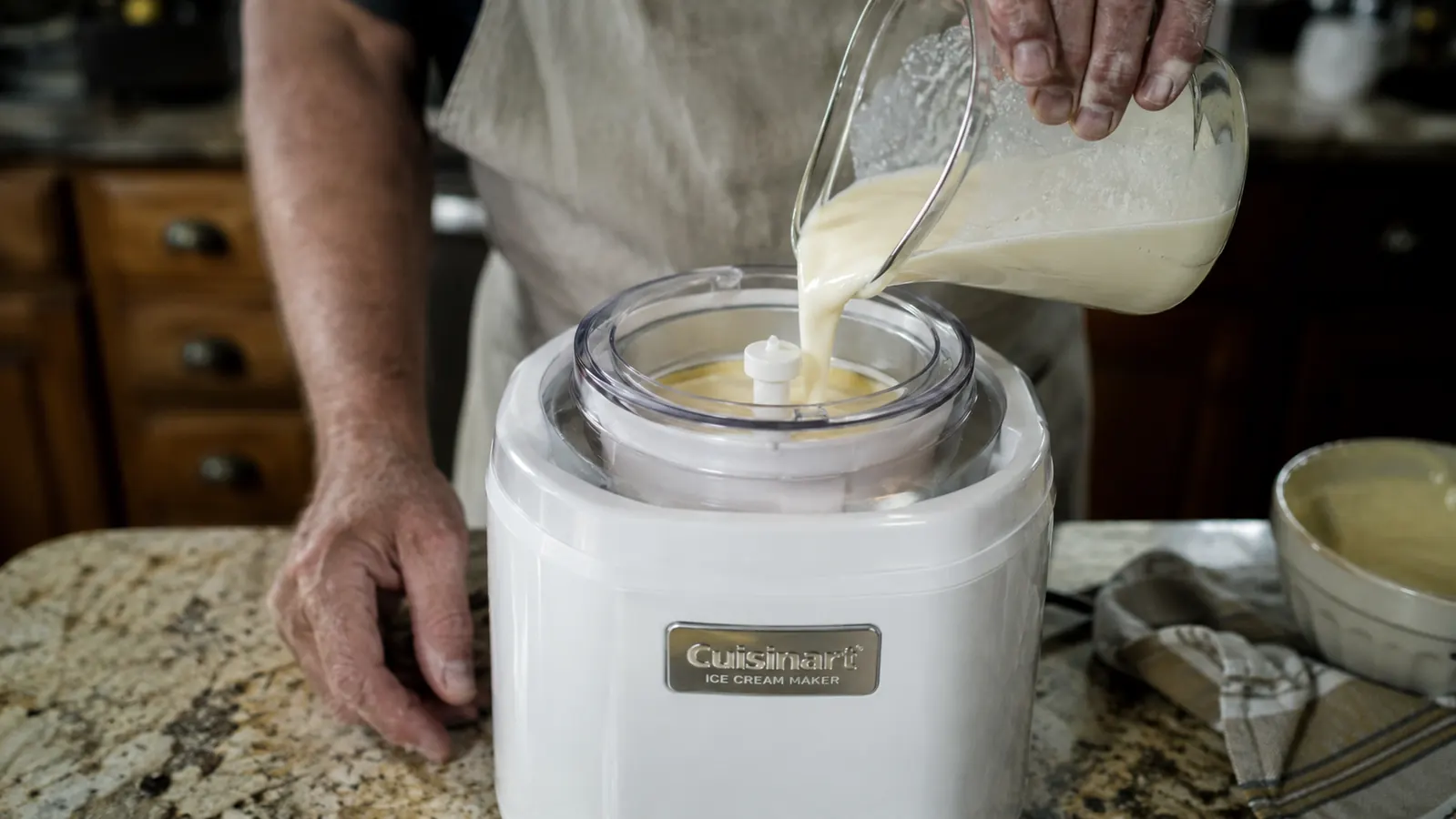 A hand pouring cream mixture into the white Cuisinart ICE-21P1 ice cream maker bowl with the paddle running