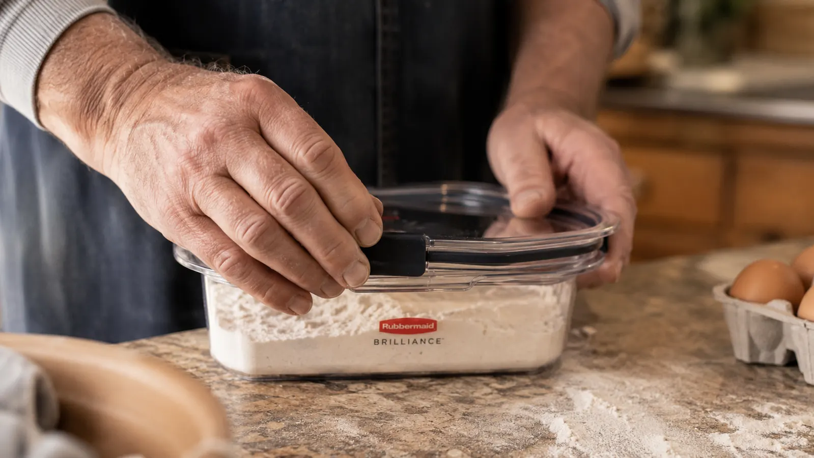 A hand snapping a latching lid onto a clear Rubbermaid Brilliance container on a kitchen counter